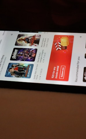 A smartphone displaying a streaming app interface, showing various movie and TV show thumbnails. The screen includes categories like 'For you' and 'New release hit films', with images from popular films. The background is a dimly lit setting with a fabric surface.