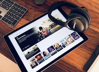 A wooden table holds a tablet displaying a streaming service interface with various movie and TV show thumbnails. Nearby, a pair of Bose headphones rests on the table along with a keyboard and a mouse.