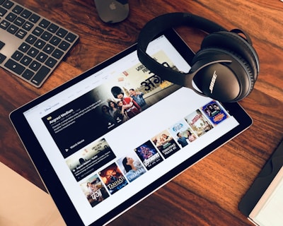 A wooden table holds a tablet displaying a streaming service interface with various movie and TV show thumbnails. Nearby, a pair of Bose headphones rests on the table along with a keyboard and a mouse.