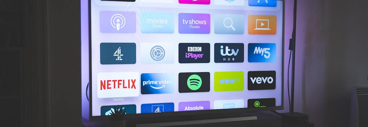 A flat-screen television is displayed in a dimly lit room, showcasing a variety of streaming service apps on its screen, such as Netflix, Prime Video, and Spotify. The TV is positioned on a black stand beside a tall floor lamp, casting a soft ambient light on the surrounding area.