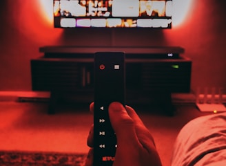 A hand holds a remote with visible buttons, pointed towards a wall-mounted TV displaying a streaming platform interface. The room is dimly lit, with a warm, red ambient light highlighting the TV screen and surroundings.