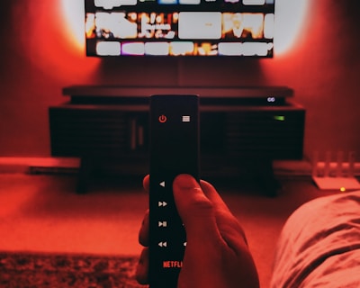 A hand holds a remote with visible buttons, pointed towards a wall-mounted TV displaying a streaming platform interface. The room is dimly lit, with a warm, red ambient light highlighting the TV screen and surroundings.