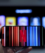 A hand holding a smartphone displaying the Netflix logo on its screen with a background of colorful vertical lines. In the background, a television screen shows blurry icons or profiles with various colors.