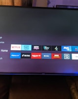 A television screen displays a smart TV interface with multiple app icons arranged in rows. Options such as Settings, Sources, Home, Search, and TV are listed on the left side. Some recognizable app icons include Netflix, Prime Video, BBC iPlayer, ITV Hub, All 4, My5, YouTube, and TikTok. The time and date are shown in the top right corner.
