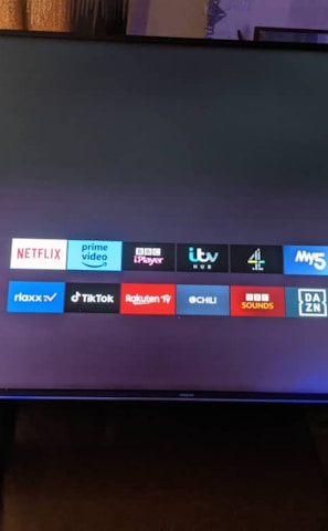 A television screen displays a smart TV interface with multiple app icons arranged in rows. Options such as Settings, Sources, Home, Search, and TV are listed on the left side. Some recognizable app icons include Netflix, Prime Video, BBC iPlayer, ITV Hub, All 4, My5, YouTube, and TikTok. The time and date are shown in the top right corner.