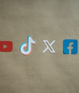 Four social media platform logos are arranged on a brown background. From left to right, the logos are for YouTube in red, TikTok in a combination of red, blue, and white, X in white, and Facebook in blue.