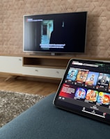 A modern living room features a flat-screen television mounted on a light-colored wooden stand with a patterned wall backdrop. Below the TV, a carpet lies on a wooden floor. In the foreground, a tablet displays a streaming platform interface with various show thumbnails, suggesting a relaxed entertainment setup.