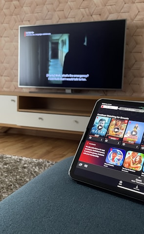 A modern living room features a flat-screen television mounted on a light-colored wooden stand with a patterned wall backdrop. Below the TV, a carpet lies on a wooden floor. In the foreground, a tablet displays a streaming platform interface with various show thumbnails, suggesting a relaxed entertainment setup.
